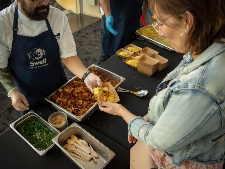 Image of A chef in an apron serves food onto a plate held by a woman wearing glasses and a denim jacket, with trays of ingredients and wooden cutlery arranged on the table.