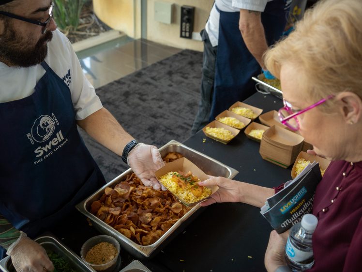 Image of A chef wearing a blue apron serves food from a tray to a woman holding a water bottle and serviette. The woman receives a dish topped with herbs, whilst more food containers sit on the table nearby.