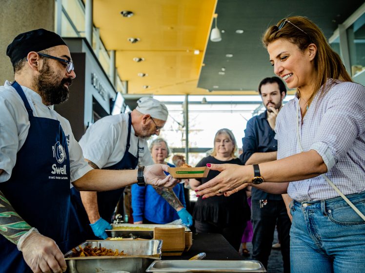 Image of A woman smiles as she receives a dish from a tattooed chef at a food stall, whilst other people queue and watch in a bright indoor market setting.