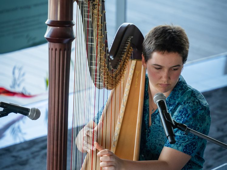 Image of A person with short hair, wearing a blue patterned shirt, plays a large wooden harp while seated on a stage. Two microphones are positioned nearby, and natural light streams in from large windows behind them.