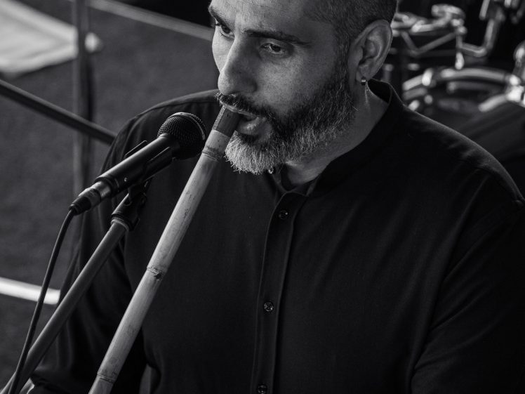 Image of A man with a shaven head and beard, dressed in a dark shirt, plays a ney flute and sings into a microphone. There is a drum kit visible in the background. The image is in black and white.