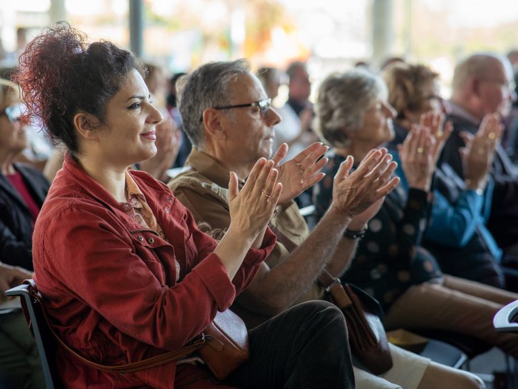 Image of A group of people sit in rows, smiling and applauding during an indoor event. The focus is on a woman in a red jacket in the front row, with others of various ages applauding beside and behind her.