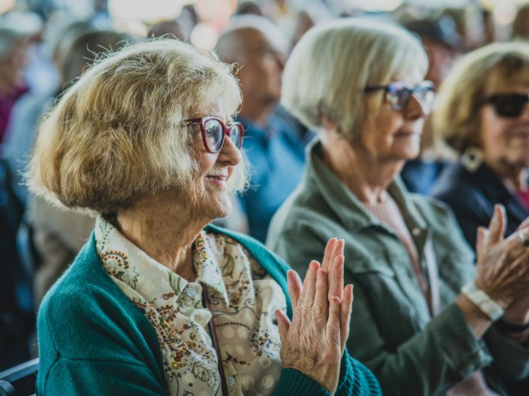 Image of Older women with glasses sit in a crowd, smiling and clapping, wearing casual clothing. The background is softly blurred, showing other people seated and applauding in what appears to be an indoor event.