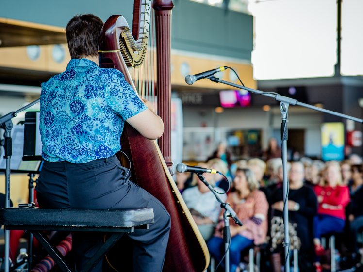 Image of A person in a blue patterned shirt plays a large harp on stage in front of microphones, with a seated audience watching in a brightly lit indoor space.