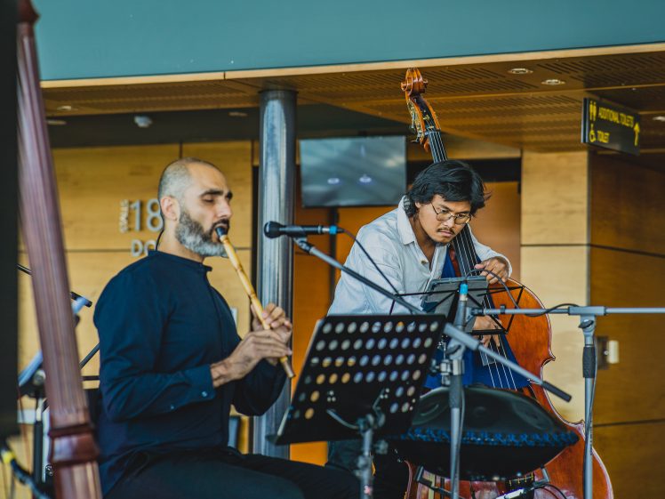 Image of Two musicians perform indoors; one plays a wooden flute, while the other plays an upright bass. They are surrounded by microphones and music stands, with a modern wooden and metal background.
