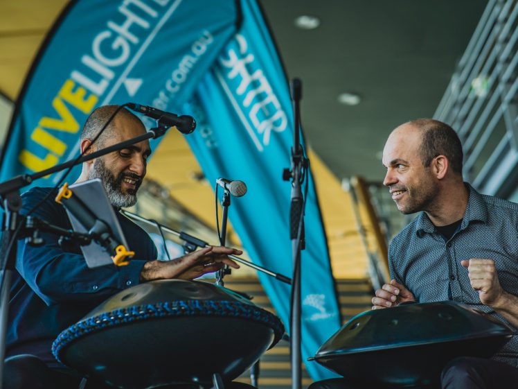 Image of Two men sit and play handpans together on stage, smiling and engaging with each other. Blue event banners and microphones are visible in the background, along with a staircase and building interior.