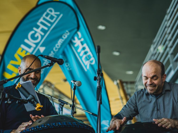 Image of Two musicians smile whilst playing hand drums on stage, surrounded by microphones. Behind them, bright blue flags display the word LiveLighter. The setting appears lively and indoors with modern architecture.