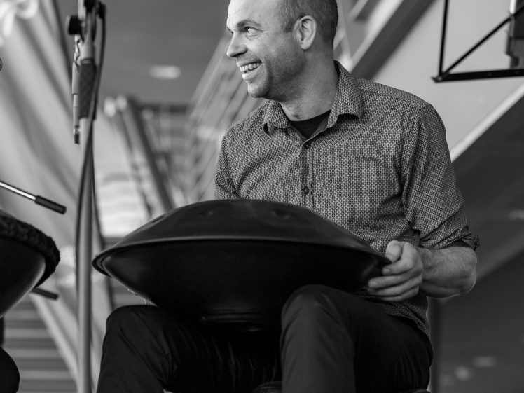 Image of A smiling man sits on a stool, playing a handpan drum. He is positioned near a microphone and music stand, with a staircase and modern interior in the background. The image is in black and white.