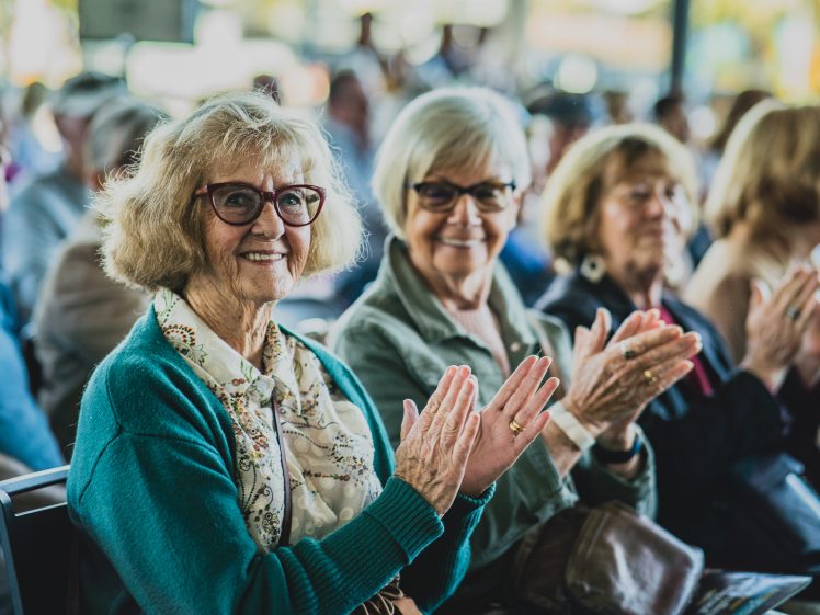 Image of Two older women wearing glasses and jumpers sit in a crowd, smiling and applauding. Other people are seated around them in the background, suggesting they are attending an event or performance.