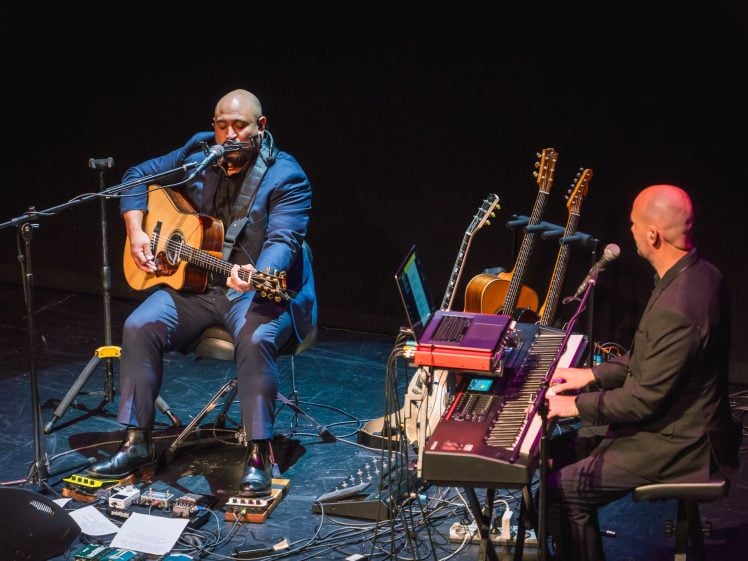 Image of Two musicians perform on stage; one sings and plays acoustic guitar with a harmonica while seated, the other plays a keyboard. Several guitars rest on stands nearby, and various audio equipment surrounds them.
