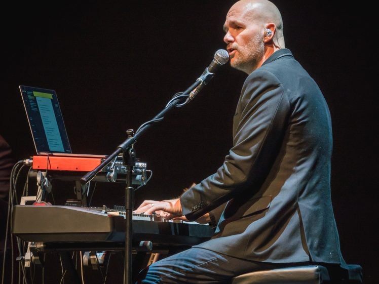 Image of A bald man in a suit plays a keyboard and sings into a microphone on stage, with a laptop and music equipment in front of him, set against a dark background.
