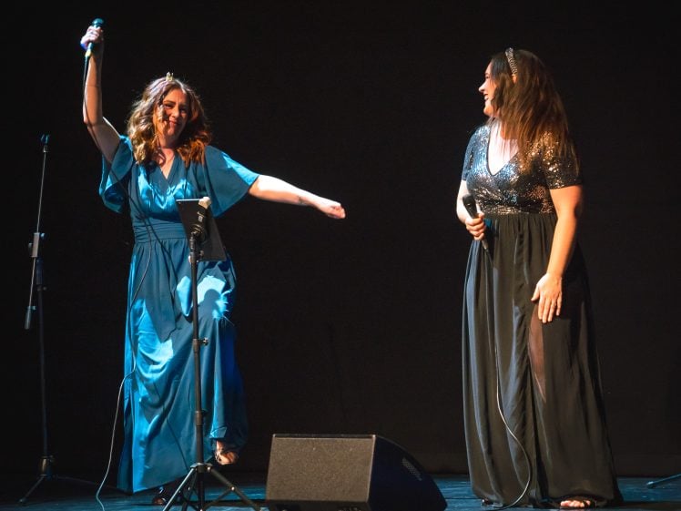 Image of Two women in elegant dresses perform on stage. One, in a blue gown, energetically lifts a microphone with one arm raised and a leg outstretched. The other, in a black dress, smiles whilst holding a microphone.