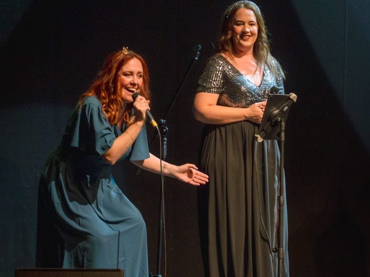 Image of Two women stand on stage under spotlights. One, in a blue dress, crouches and gestures with a microphone, while the other, in a sparkly silver top and black skirt, smiles and stands at a microphone stand holding a tablet.