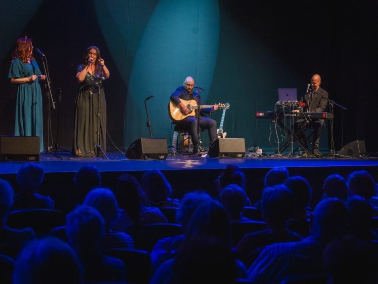 Image of Four musicians perform on stage: two women sing at microphones, a man plays acoustic guitar and sings, and another man plays keyboard. Blue stage lighting highlights the performers, and an audience watches in the foreground.