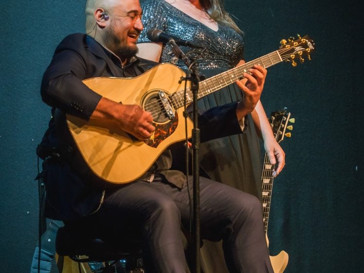 Image of A man sitting and playing an acoustic guitar while smiling, with a woman standing behind him, also smiling and holding part of the guitar. Both are dressed in formal attire on a dimly lit stage.