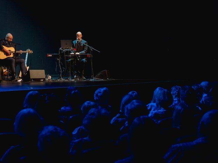 Image of Two musicians perform on a dimly lit stage, one playing an acoustic guitar and the other on a keyboard, while an audience watches from the darkened theatre. Blue lighting creates a calm atmosphere.