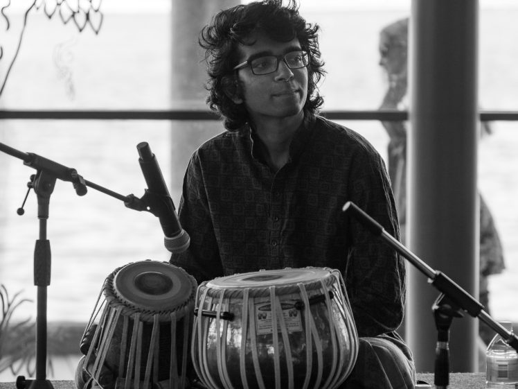 Image of A person wearing glasses sits on stage playing a pair of tabla drums, surrounded by microphones. The background shows an outdoor setting with blurred trees and water. The image is in black and white.