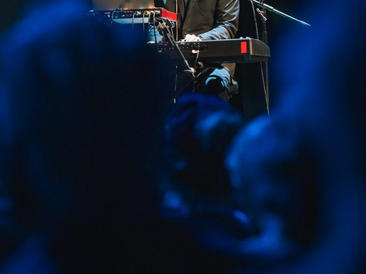 Image of A bald man in a suit plays keyboard and sings into a microphone on stage, with a laptop in front of him. The foreground shows blurred audience members under blue lighting.