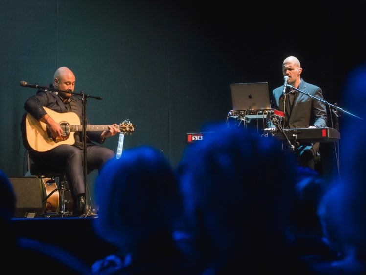 Image of Two bald men in suits perform on stage; one plays an acoustic guitar and sings into a microphone, while the other plays a keyboard and sings. An audience watches them in a dimly lit setting.