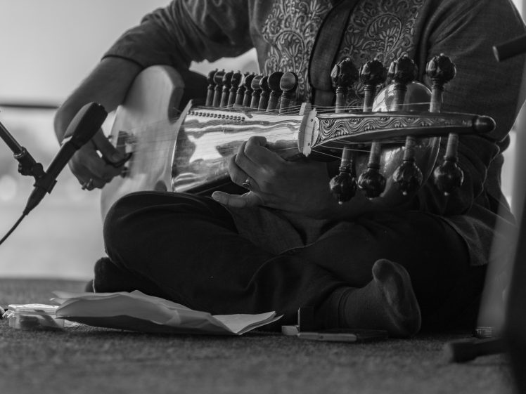 Image of A person sits cross-legged on the floor, playing a traditional stringed instrument called a sarod. The image is in black and white, focusing on their hands and the instrument, with a microphone nearby.