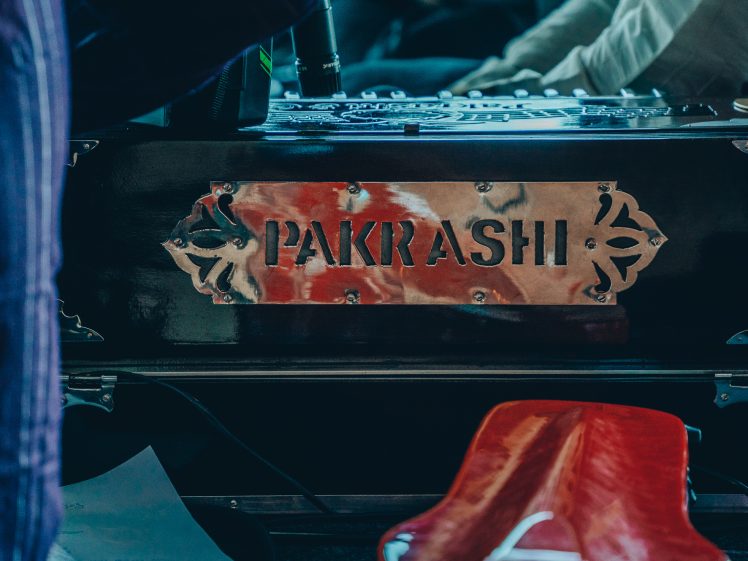 Image of Close-up of a harmonium with a metallic label reading Pakrashi on its front panel; part of a red object and some papers are visible in the foreground.