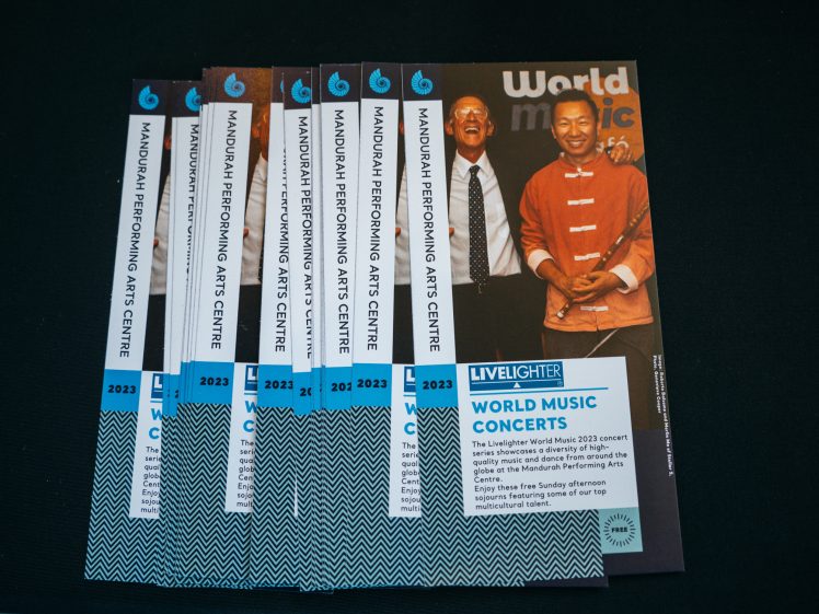 Image of A stack of leaflets for World Music Concerts at Mandurah Performing Arts Centre, featuring a smiling older man and woman on the cover, with blue and white event details and 2023 printed along the left edge.