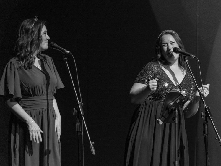 Image of Two women in dresses sing into microphones on stage. One smiles broadly and gestures with her hands, while the other looks at her with a pleasant expression. The scene is lit dramatically, and the background is dark.