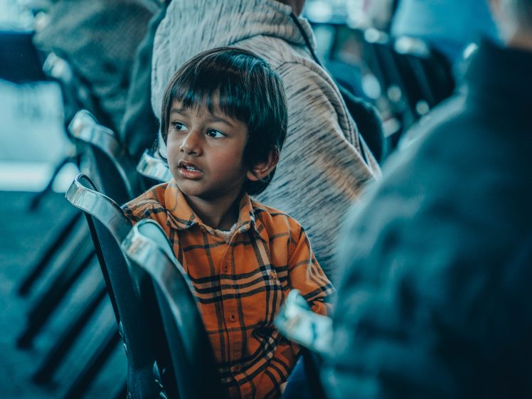 Image of A young child in an orange checked shirt sits on a chair, looking to the side with a curious expression. Other people are seated nearby, but their faces are out of focus.