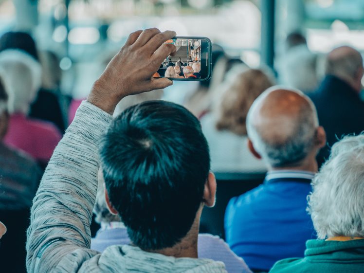 Image of A person in a grey hoodie holds up a mobile phone to take a photo or video of an event, surrounded by a seated audience with their backs to the camera.