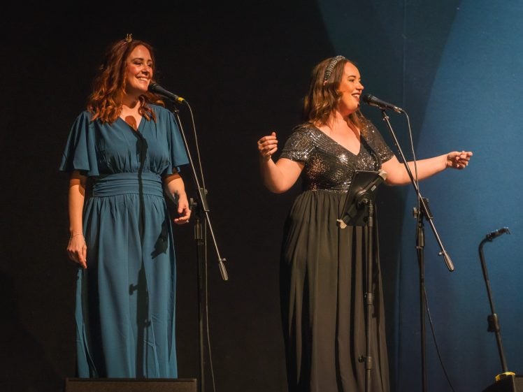 Image of Two women in long dresses sing into microphones on stage. One wears a blue dress and the other a sparkly black dress. Both are smiling under stage lights against a dark background.