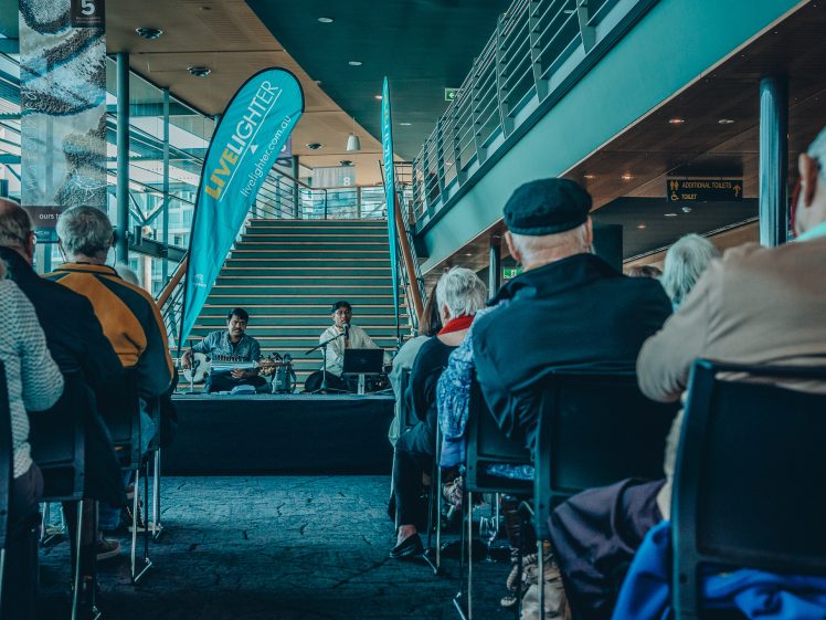 Image of Audience members sit facing a small stage where two musicians perform indoors near a staircase. A blue banner with “LIVELIGHTER” is displayed beside the stage. The setting has modern architecture and large windows.