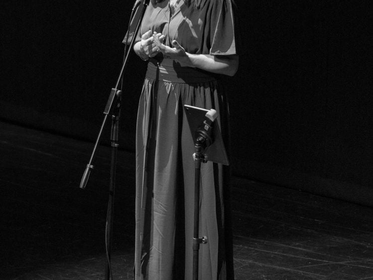 Image of A woman in a long dress stands on stage, speaking into a microphone. She is lit by a spotlight, with a dark background behind her. The image is in black and white.