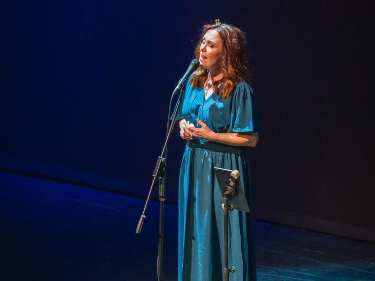 Image of A woman in a long blue dress stands alone on a dimly lit stage, singing into a microphone with a music stand nearby.