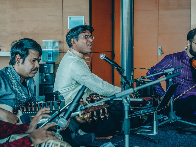 Image of Four musicians perform indoors. One sings into a microphone, one plays a string instrument, one plays a tabla drum, and another plays a violin. They wear traditional South Asian attire and sit surrounded by musical equipment.