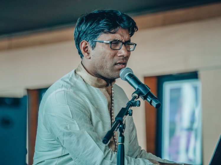 Image of A man wearing glasses and a traditional light-coloured kurta sings or speaks into a microphone whilst playing a keyboard indoors.
