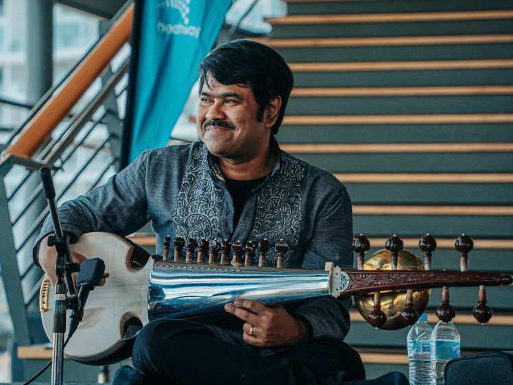 Image of A man in a patterned shirt sits cross-legged, smiling whilst holding a traditional string instrument. A microphone and two water bottles are nearby, and a staircase is in the background.