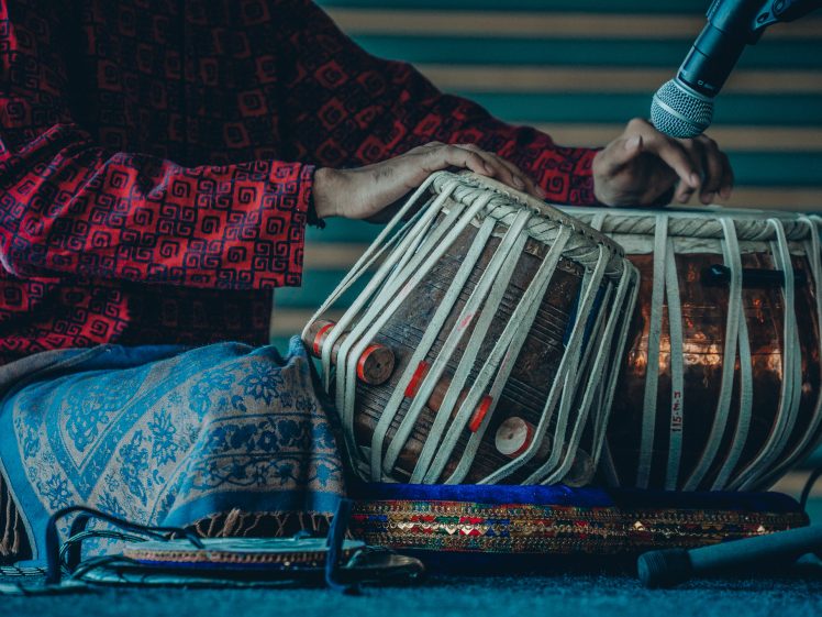 Image of A person in a red patterned shirt plays the tabla, a pair of traditional Indian drums, with a microphone positioned nearby to capture the sound.