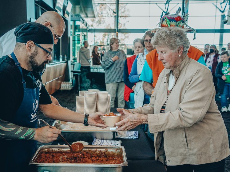 Image of A man wearing an apron and gloves serves food from a tray into a bowl held by an elderly woman in a busy indoor setting, with a queue of people waiting in the background.