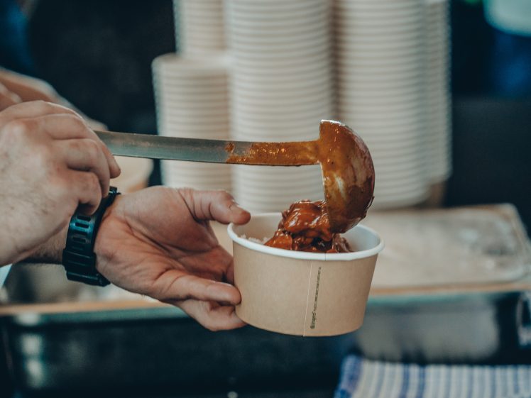 Image of A person wearing a watch ladles stew or curry into a brown paper bowl, with stacks of disposable cups in the background.