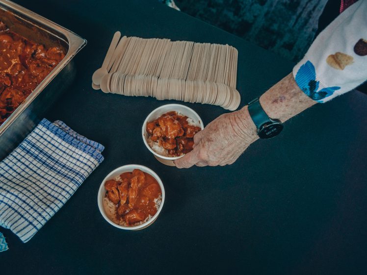 Image of A person wearing a long-sleeve shirt and watch serves a bowl of stew next to another filled bowl, a tray of food, a stack of wooden spoons, and a striped tea towel on a dark table.