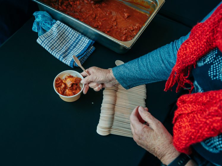 Image of A person wearing a red scarf holds a wooden spoon near a cup of food, with a tray of red sauce and a stack of wooden cutlery on a dark table nearby.