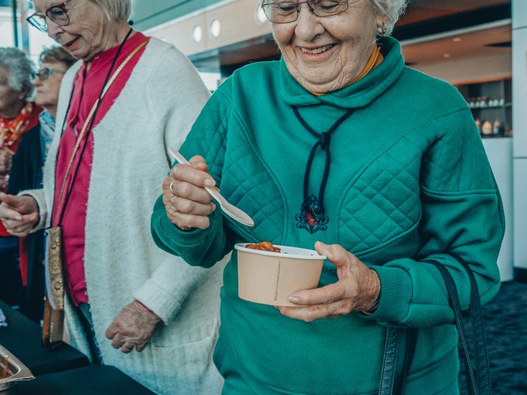 Image of An elderly woman in a green jumper smiles whilst holding a bowl of food and a spoon. She stands beside another older woman in a pink top and white cardigan at a buffet table indoors.