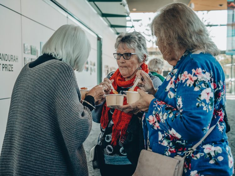 Image of Three older women stand together indoors, eating from bowls and talking. One wears a red scarf, another a blue floral top, and the third a grey shawl. Art is displayed on the wall behind them.