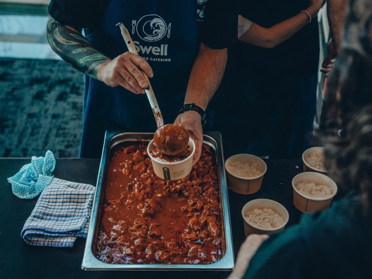 Image of A person in a blue apron serves a scoop of stew from a large tray into a paper bowl. Nearby, bowls of white rice are ready to be served. Other people stand nearby, partially visible.