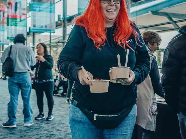 Image of A person with long bright red hair, glasses, and a black hoodie smiles while holding two bowls of food in a modern, sunlit indoor space with other people and large banners in the background.