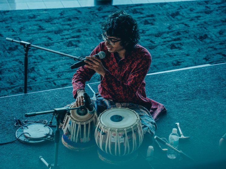 Image of A person wearing glasses and a patterned red shirt plays tabla drums and sings into a microphone on stage, with microphones and instruments around them.