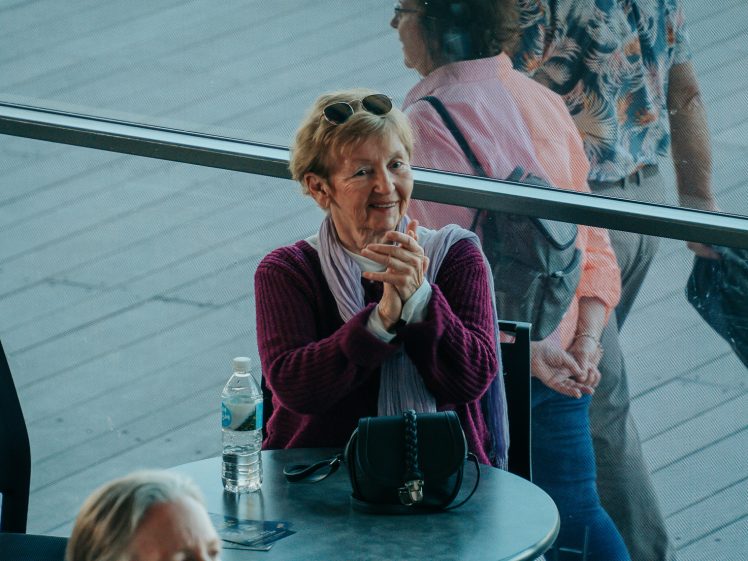 Image of An older woman with sunglasses on her head sits at a table, smiling and clasping her hands. A water bottle and handbag are on the table. Two people walk past in the background, seen through glass.