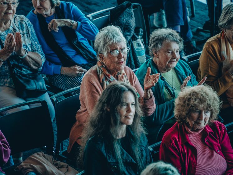Image of A group of older women sit closely together in rows of chairs, some clapping and others smiling, appearing to enjoy a live event or performance. The atmosphere is lively and friendly.