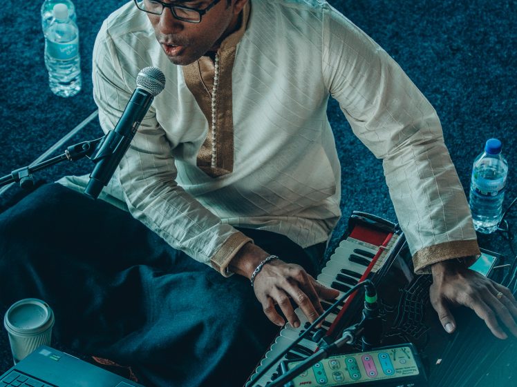Image of A man in traditional attire sings into a microphone while playing a harmonium. He sits on a dark carpet, surrounded by a laptop, a small device, and water bottles.