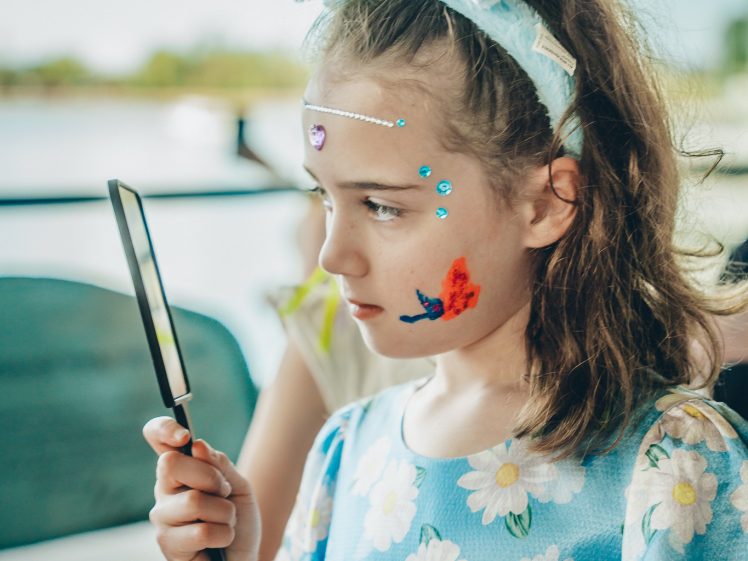 Image of A young girl wearing blue bunny ears and a floral dress looks into a handheld mirror. She has face paint, including a butterfly on her cheek and gems on her forehead, standing indoors near a window.